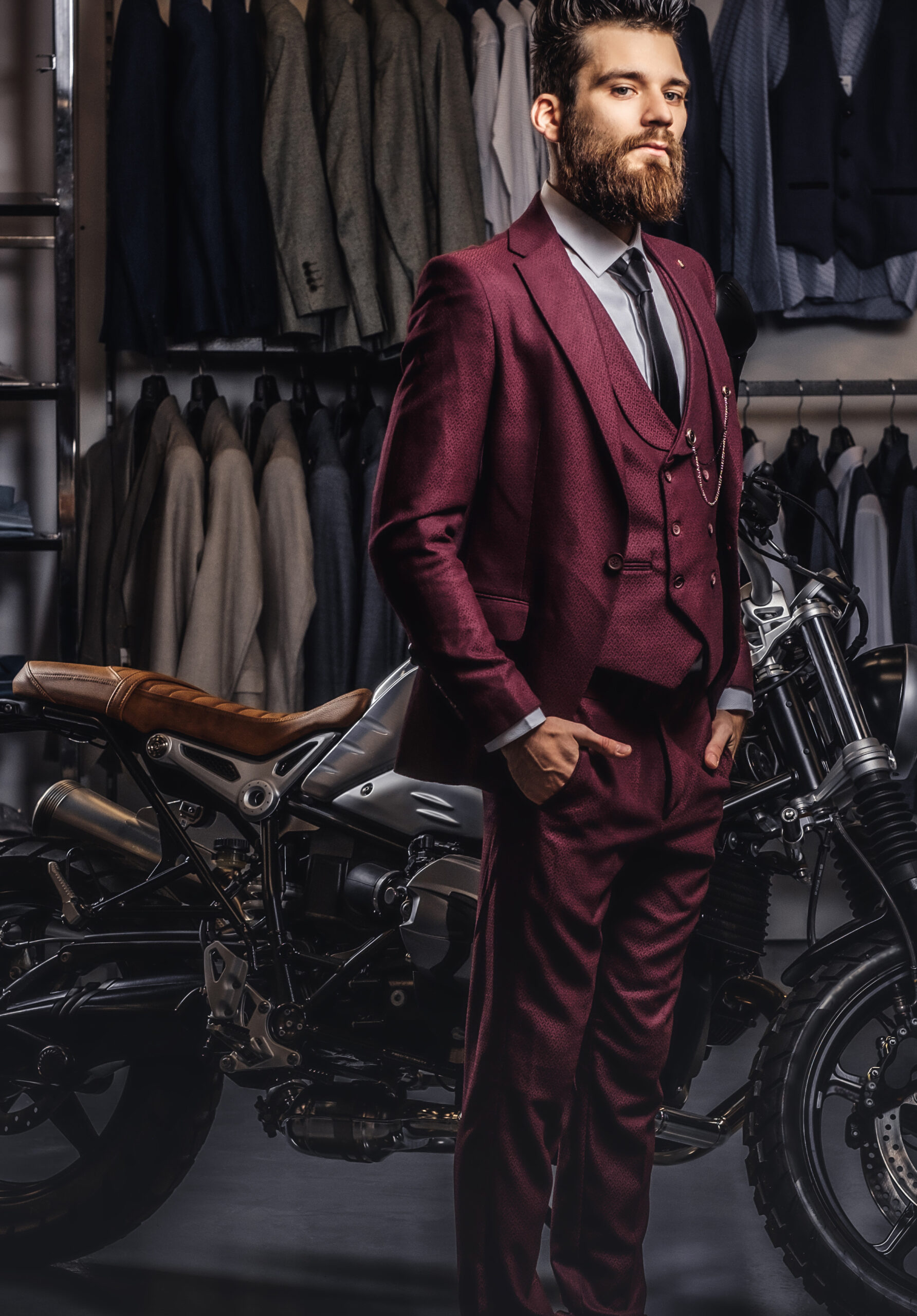Stylish handsome man dressed in vintage red suit posing  with hands in pockets near retro sports motorbike at men's clothing store.